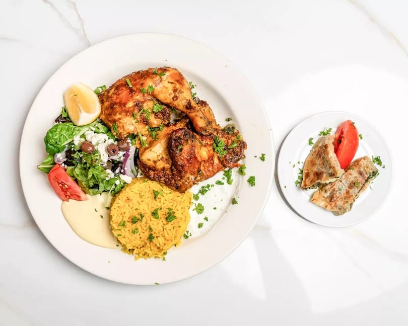 A plate of roasted chicken, yellow rice, Greek salad, and a side of spinach pie.