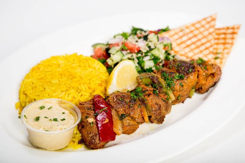 A plate of beef shish kebab with yellow rice, tabbouleh salad, hummus, and pita bread.