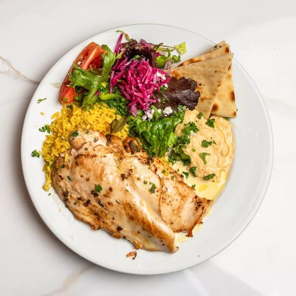 Grilled chicken breast with yellow rice, hummus, pita bread, and a fresh salad.